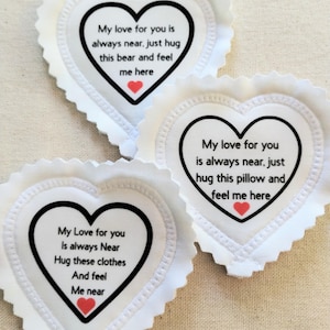 May include: Three white heart-shaped fabric patches with pinked edges. Each patch features a black heart outline, a small red heart icon, and printed text. The text reads: My love for you is always near, just hug this bear/pillow/clothes and feel me here.