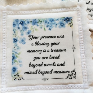 May include: White fabric square with a scalloped edge, featuring a floral design in shades of blue. The text reads: "Your presence was a blessing, your memory is a treasure, you are loved beyond words and missed beyond measure."
