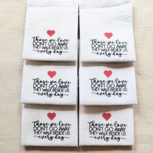 May include: Six white rectangular fabric labels arranged in two columns. Each label features a small red heart icon above the black printed text: Those we love DON'T GO AWAY THEY WALK BESIDE US every day, written in a mix of script and sans-serif fonts.