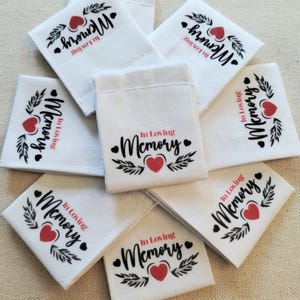 May include: A collection of white fabric labels featuring the black text In Loving Memory, a red heart icon, and black leaf illustrations. Each rectangular label is oriented differently, showcasing the repeated design printed on the white material.