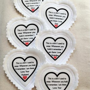 May include: White heart-shaped fabric patches with scalloped edges. Each patch has a black border and contains text in black font with a red heart. The text reads, "This is a shirt I used to wear. Whenever you hold it remember I am there. Love, Mom/Dad/Grandma/Grandpa/Your Husband/Your Wife."