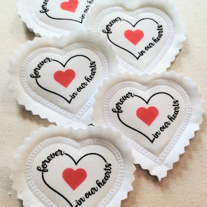 May include: Five white heart-shaped fabric patches with scalloped edges. Each patch features a central red heart graphic surrounded by the black text "forever in our hearts" written in a cursive script that forms a larger heart outline.
