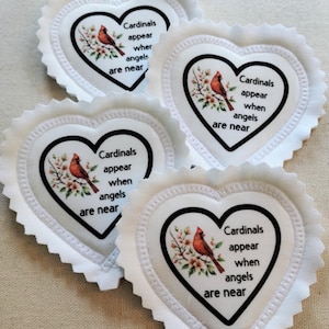 May include: Four heart-shaped fabric ornaments with pinked edges. Each features a printed illustration of a red cardinal bird perched on a flowering branch and the black text: Cardinals appear when angels are near, enclosed in a black heart outline.