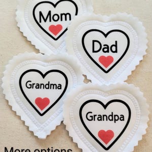 May include: Four white heart-shaped coasters with black outlines and scalloped edges. Each coaster has a name in black text: Mom, Dad, Grandma, and Grandpa. A red heart is inside each heart shape. The words "More options" are at the bottom.