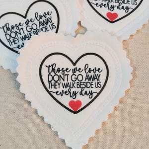 May include: A white heart-shaped fabric patch with a scalloped edge. The patch features black text that reads, "Those we love DON'T GO AWAY THEY WALK BESIDE US every day," and a small red heart icon positioned below the text.