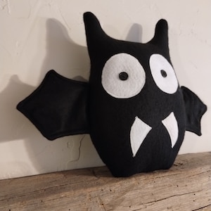 May include: Black felt bat plush toy with white eyes and fangs. The toy has a stitched-on smile and is sitting on a wooden surface.