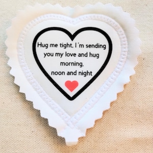 May include: A white heart-shaped fabric patch with a scalloped edge. The center features a black heart outline containing the text: Hug me tight, I'm sending you my love and hug morning, noon and night, with a small red heart icon below the text.
