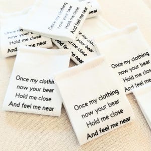 May include: A collection of small, white fabric tags with black text. Each tag has the phrase "Once my clothing, now your bear, Hold me close, And feel me near" printed on it. The tags are scattered on a light-colored surface.