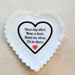 May include: A white heart-shaped fabric patch with a scalloped edge. The center features a black heart outline containing the black text: Once my shirt, Now a bear, Hold me close, I'll be there, with a small red heart printed below the text.