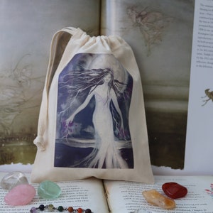Puede incluir: Una bolsa de tela blanca con una imagen impresa de una mujer de pie en un bosque iluminado por la luna. La bolsa est&aacute; llena de cristales de colores y una cadena de plata con cuentas.