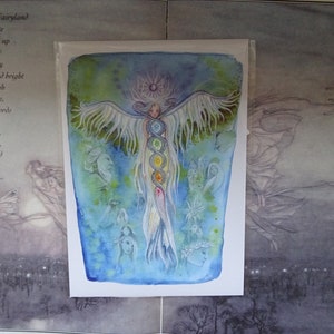 May include: A watercolor painting of a winged figure with a halo, surrounded by smaller figures. The central figure has a long, flowing tail and is adorned with seven colorful circles, possibly representing chakras. The background is a soft blue with hints of green.