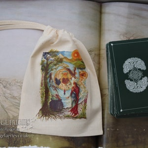 May include: A white cloth bag with a drawstring closure. The bag has a colorful illustration of a fairy scene with a cauldron, a tree, and a woman in a red dress. The bag is sitting on a table next to a stack of green tarot cards with a white design. The text on the bag reads "Free Range Fairies artwork by Kate Monkman www.freerangefairies.co.uk"