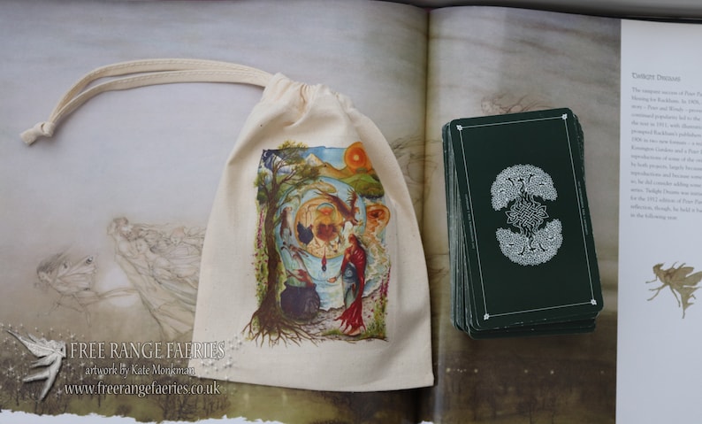 May include: A white cloth bag with a drawstring closure. The bag has a colorful illustration of a fairy scene with a cauldron, a tree, and a woman in a red dress. The bag is sitting on a table next to a stack of green tarot cards with a white design. The text on the bag reads "Free Range Fairies artwork by Kate Monkman www.freerangefairies.co.uk"