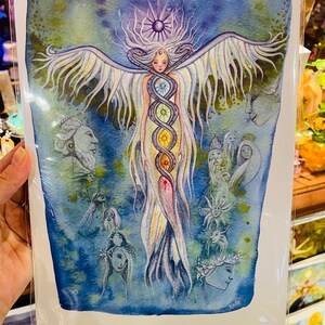 Puede incluir: Pintura acuarela de una mujer con alas y una serpiente de colores arco&iacute;ris envuelta alrededor de su cuerpo. La mujer est&aacute; rodeada de otras figuras y un cielo azul con estrellas.