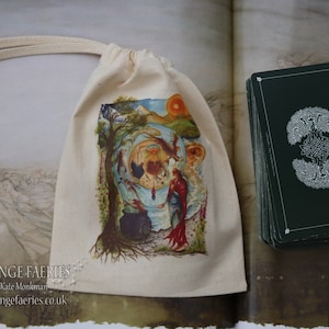May include: A white canvas bag with a drawstring closure. The bag features a colorful illustration of a woman standing in a forest with a cauldron and a tree. The bag has the text "FREE RANGE FAERIES" printed on the front. The bag is sitting on top of a book with a deck of cards.