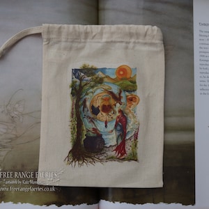 May include: A white cotton drawstring bag with a colorful illustration of a fairy scene. The illustration features a fairy, a cauldron, a tree, and a red sunset. The bag has the text "FREE RANGE FAERIES" and "artwerk by Kate Monkman" printed on it.