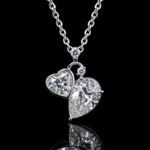 Puede incluir: Un collar de tono plateado con un colgante que presenta dos piedras transparentes facetadas, una en forma de corazón y otra en forma de pera, suspendidas de una cadena fina. Dos piedras pequeñas, redondas y transparentes coronan el colgante.