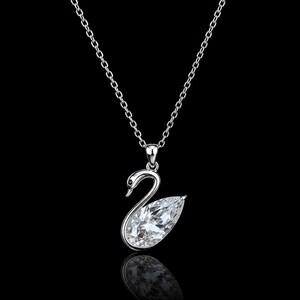 Puede incluir: Un collar de tono plateado con un colgante en forma de cisne. El cuerpo del cisne está formado por una gran piedra preciosa transparente tallada en forma de pera, mientras que el cuello y la cabeza son de metal pulido con un pequeño detalle negro para el ojo.
