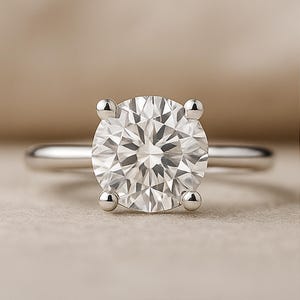 Könnte beinhalten: Nahaufnahme eines Solitär-Verlobungsrings. Der Ring hat einen großen, runden Diamanten im Brillantschliff, der in einer Vier-Krappen-Fassung gefasst ist. Das Band ist schlicht und poliert silberfarben.