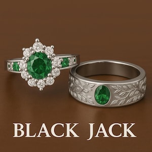 Può includere: Due anelli con pietre preziose verde smeraldo. Uno presenta un grande smeraldo rotondo circondato da diamanti più piccoli. L'altro ha uno smeraldo ovale e un motivo a foglie. Il testo "BLACK JACK" è in basso.