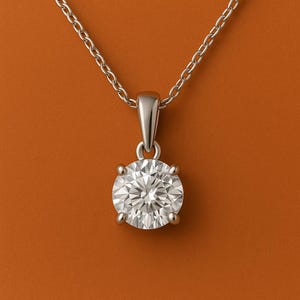 Op de afbeelding: Een zilveren hangerketting met een grote, ronde, briljant geslepen diamant. De diamant wordt vastgehouden in een vier-klauwzetting en hangt aan een delicate ketting. De ketting wordt gepresenteerd tegen een warme, oranje-bruine achtergrond.