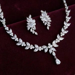 May include: A sparkling silver necklace and matching earrings set. The jewelry features a floral design with clear, brilliant-cut stones. The necklace has a teardrop pendant. The set is displayed on a dark burgundy background.