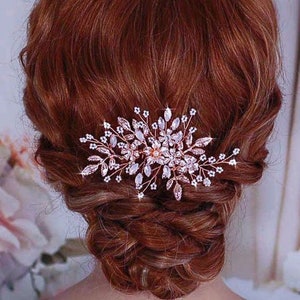 May include: A rose gold hair comb with clear crystals and small white flowers. The comb is in the hair of a person with auburn hair, styled in a low bun. The comb is a decorative hair accessory.