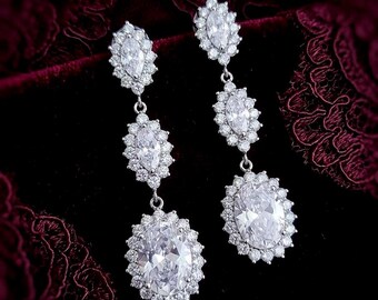 May include: A pair of silver-toned drop earrings featuring three vertically aligned clear stones. Each stone is surrounded by a halo of small round clear stones. The top two stones are marquise-shaped, while the bottom stone is oval-shaped.