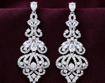 May include: A pair of silver-toned chandelier earrings featuring a teardrop-shaped stone at the top, followed by a series of intricate, scrolling metalwork sections encrusted with numerous small, clear, round-cut and pear-shaped cubic zirconia stones.