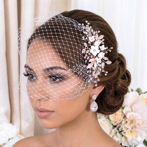 May include: A bridal headpiece with a white birdcage veil, a floral hair comb with white flowers, and sparkling crystals. The comb has rose gold leaves and accents. The bride is also wearing teardrop earrings.