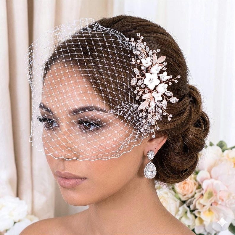 May include: A bridal headpiece with a white birdcage veil, a floral hair comb with white flowers, and sparkling crystals. The comb has rose gold leaves and accents. The bride is also wearing teardrop earrings.