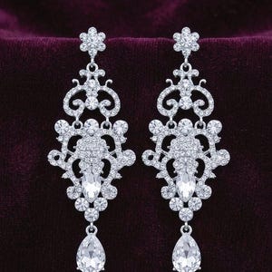 May include: A pair of silver-toned chandelier earrings featuring a floral stud top, an intricate filigree frame encrusted with numerous small clear round crystals, a central marquise-cut crystal, and a dangling pear-shaped crystal drop at the base.