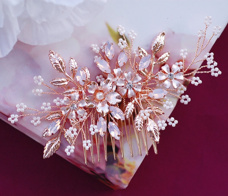 May include: A rose gold hair comb with floral and leaf detailing. The comb features clear and pink crystals, small white beads, and delicate metal leaves. Ideal for weddings or special occasions.