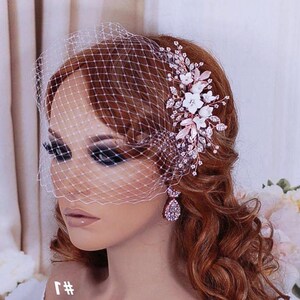 May include: A bridal headpiece with a white net veil and a floral hair clip. The clip features white flowers, pink leaves, and sparkling crystals. Teardrop earrings with crystal accents are also visible. The hair is auburn.