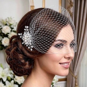 May include: A white birdcage veil featuring a delicate diamond-patterned netting. The veil is accented with a decorative cluster of clear crystals and small white pearls, positioned to the side of the head to create a classic, elegant look.
