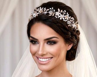 May include: A gold-toned bridal headpiece featuring a delicate arrangement of metallic leaves and small white floral accents encrusted with clear crystals. The headpiece is worn across the crown of the head, paired with a sheer white veil.