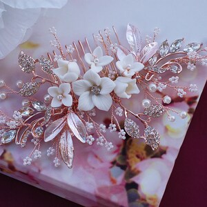 May include: A rose gold hair comb decorated with white porcelain flowers, clear crystals, and faux pearls. The comb features delicate leaves and sprigs, creating an elegant, floral design. Ideal for weddings or special occasions.