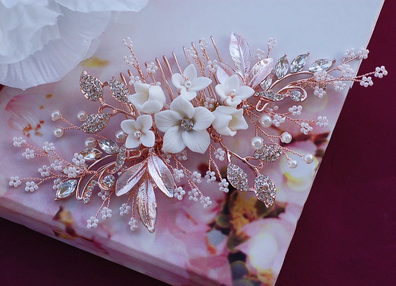 May include: A rose gold hair comb decorated with white porcelain flowers, clear crystals, and faux pearls. The comb features delicate leaves and sprigs, creating an elegant, floral design. Ideal for weddings or special occasions.