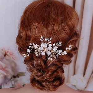 May include: A gold-coloured hair comb adorned with white fabric flowers, clear crystals, and fine sprigs. The comb has a floral design and is set against auburn hair, suggesting a bridal or special occasion accessory.