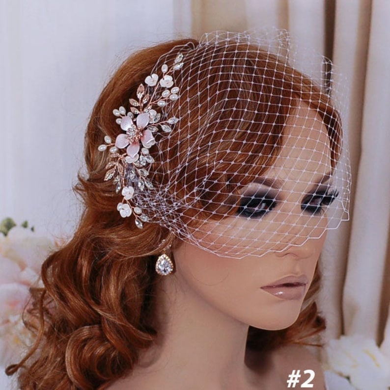 May include: A bridal headpiece with a white birdcage veil and a floral hair comb. The comb features pink flowers, crystals, and pearls. This headpiece is designed for a wedding or special occasion.