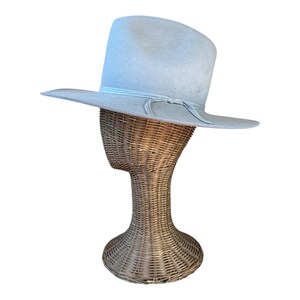Pode incluir: Um chapéu estilo fedora azul claro com aba larga e uma faixa de fita combinando amarrada em um laço. O chapéu é exibido em uma cabeça de manequim de vime. A aba do chapéu tem aproximadamente 9 cm de largura.