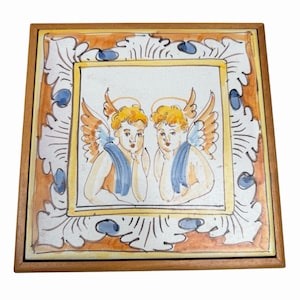 May include: A square ceramic tile framed in wood, featuring a hand-painted illustration of two winged cherubs with golden hair and blue sashes. The border displays an ornate white and orange scrollwork pattern with small blue accents.