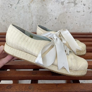 Könnte beinhalten: Ein Paar weiße Lederschuhe mit einem weißen Satinband, das um die Knöchel gebunden ist. Die Schuhe haben ein strukturiertes Obermaterial aus Leder und eine weiße Gummisohle.