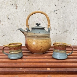 May include: A ceramic teapot with a bamboo handle and two matching mugs. The teapot and mugs are decorated with a blue, yellow, and brown glaze.