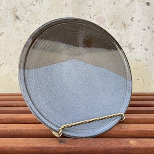 Puede incluir: Un plato de cerámica con un esmalte marrón y blanco. El plato tiene un borde ligeramente elevado y está sentado en un soporte de madera.