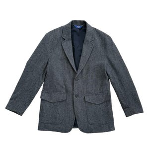 May include: A gray tweed blazer with a two-button closure and two front pockets. The blazer has a notched lapel and a dark blue interior lining. The sleeves are long and the overall style is classic and tailored.