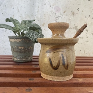 Puede incluir: Un tarro de miel de cerámica marrón con un cucharón de madera. El tarro tiene una tapa con un pomo y está decorado con pinceladas marrones y negras. El tarro está sobre una superficie de madera.