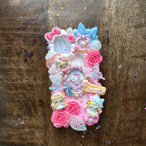 May include: A pink phone case decorated with Hello Kitty, bows, roses, and figurines. The case has a frame with a Hello Kitty image and the word "Lovely" in yellow. The case is embellished with white, pink, and blue accents.