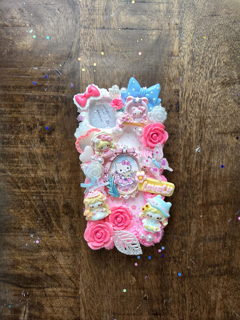 May include: A pink phone case decorated with Hello Kitty, bows, roses, and figurines. The case has a frame with a Hello Kitty image and the word "Lovely" in yellow. The case is embellished with white, pink, and blue accents.