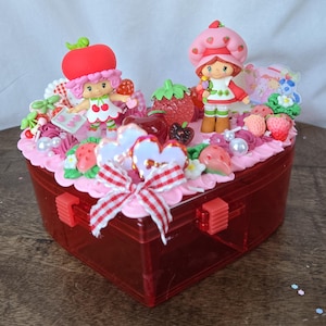 Może przedstawiać: Czerwone, plastikowe pudełko w kształcie serca, ozdobione figurkami Strawberry Shortcake, truskawkami i kwiatowymi akcentami. Pudełko posiada czerwoną i białą kratkowaną kokardę i jest wypełnione różowym lukrem i perłami.